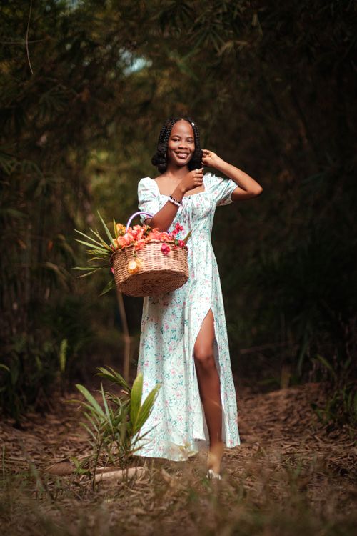 Free Cheerful woman in floral dress carrying a wicker basket filled with flowers and fruits outdoors. Stock Photo