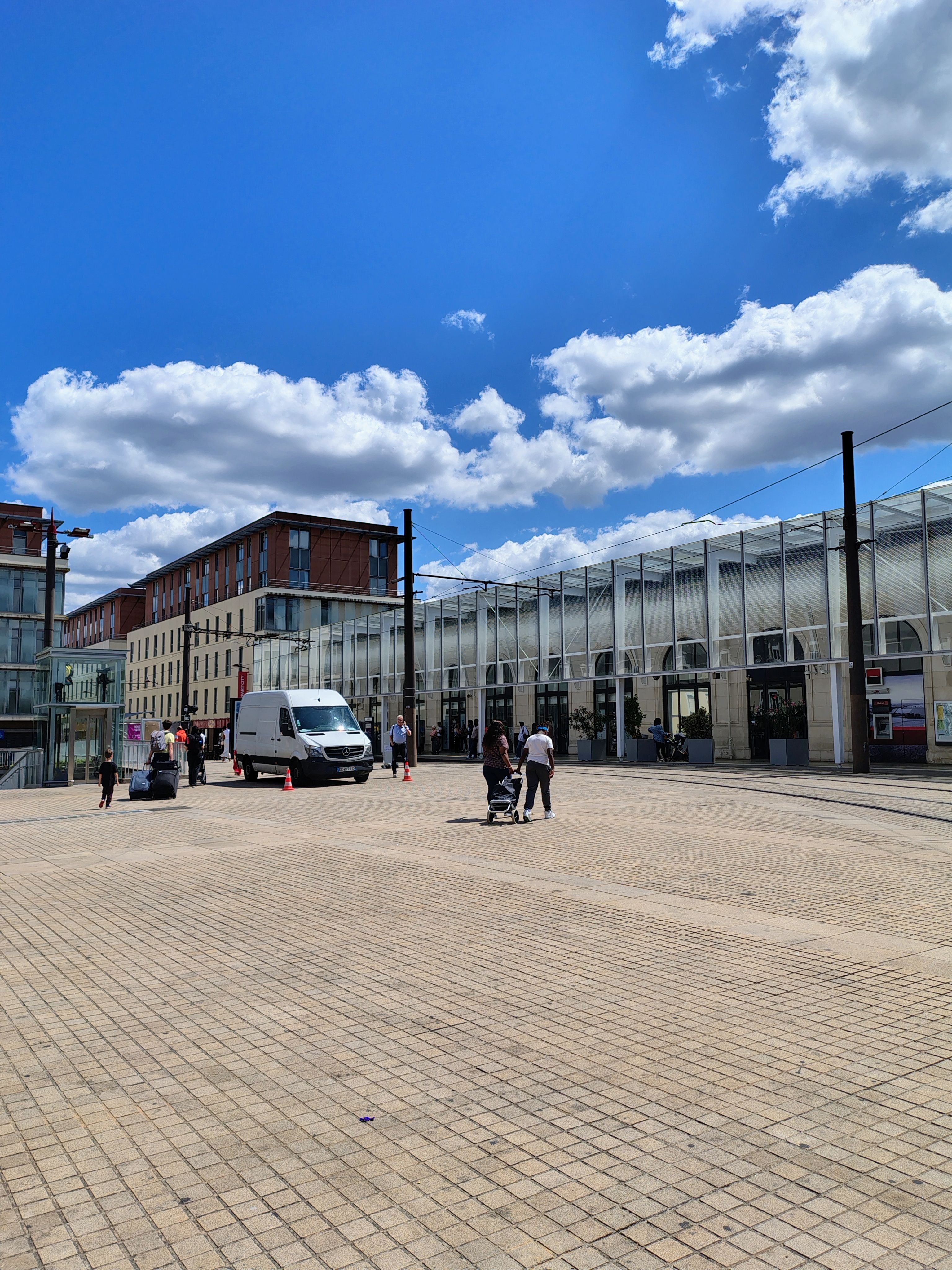 A broad paved civic square with a long glazed public building down one side, tram rails in the paving and small groups of people crossing on foot.