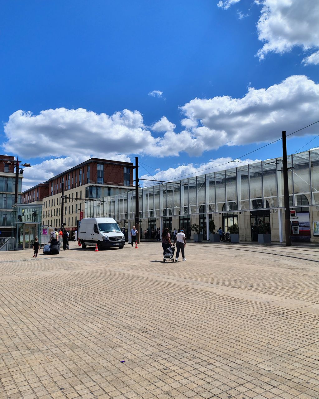 A broad paved civic square with a long glazed public building down one side, tram rails in the paving and small groups of people crossing on foot.