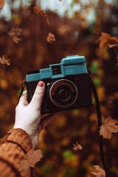 A hand holding a vintage camera surrounded by falling autumn leaves.