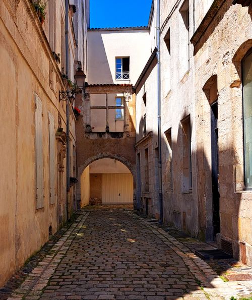 Free Charming narrow cobblestone street in a European town, framed by historic buildings and a stone archway, under a clear blue sky. Stock Photo