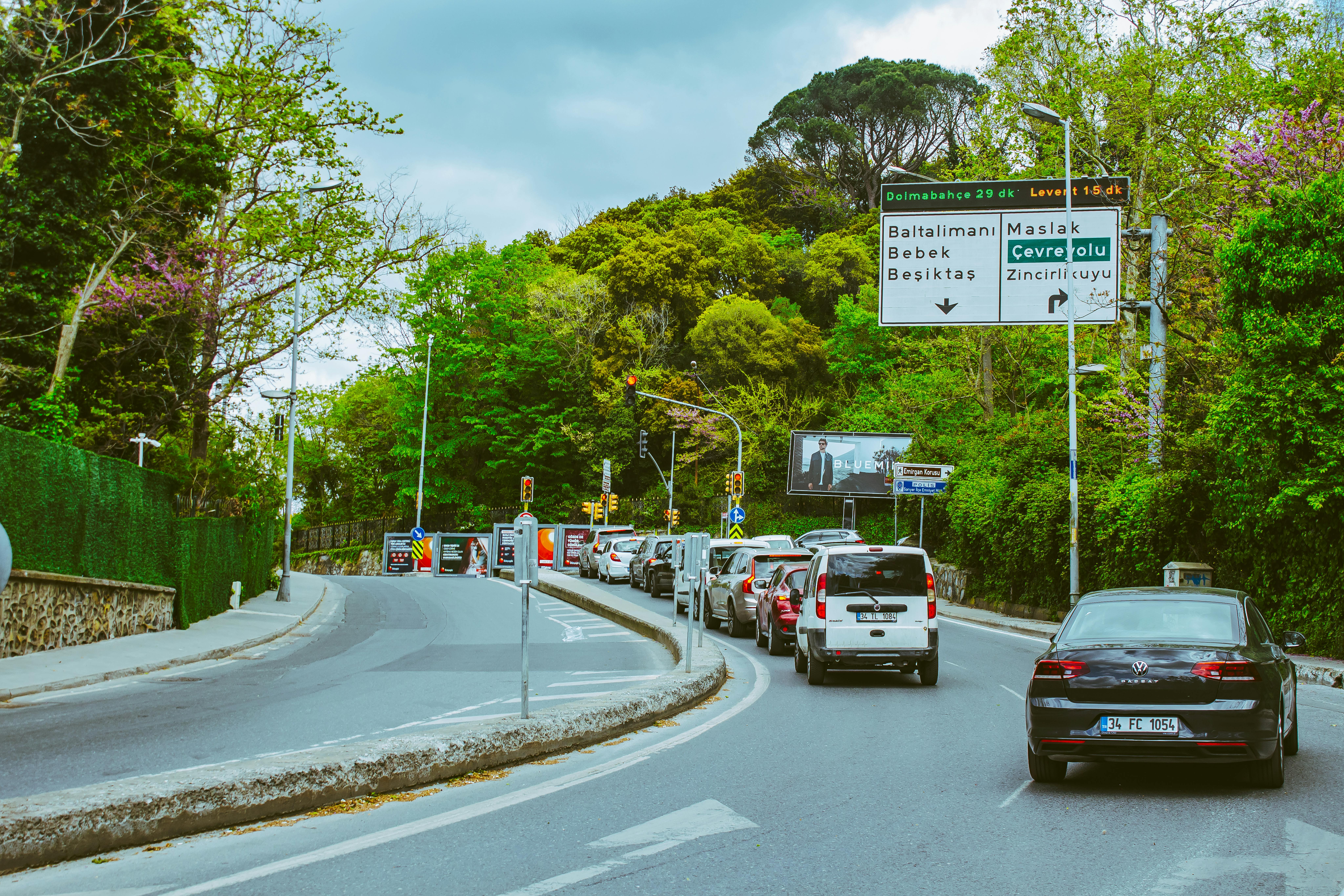 Cars line up on a lush green road under a cloudy sky, with prominent road signs.