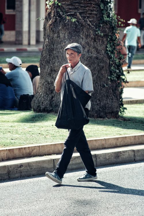 Free An elderly man strolls past outdoor seating on a sunny day in an urban setting. Stock Photo