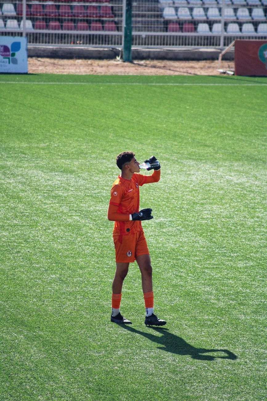 Een voetbalkeeper in oranje tenue neemt een waterpauze tijdens een zonnige wedstrijd op een groen veld.