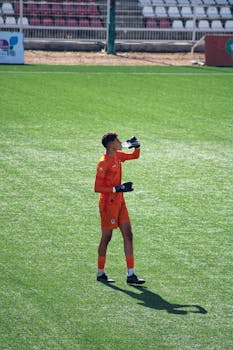 A soccer goalkeeper in orange attire takes a water break during a sunny day match on a green field.
