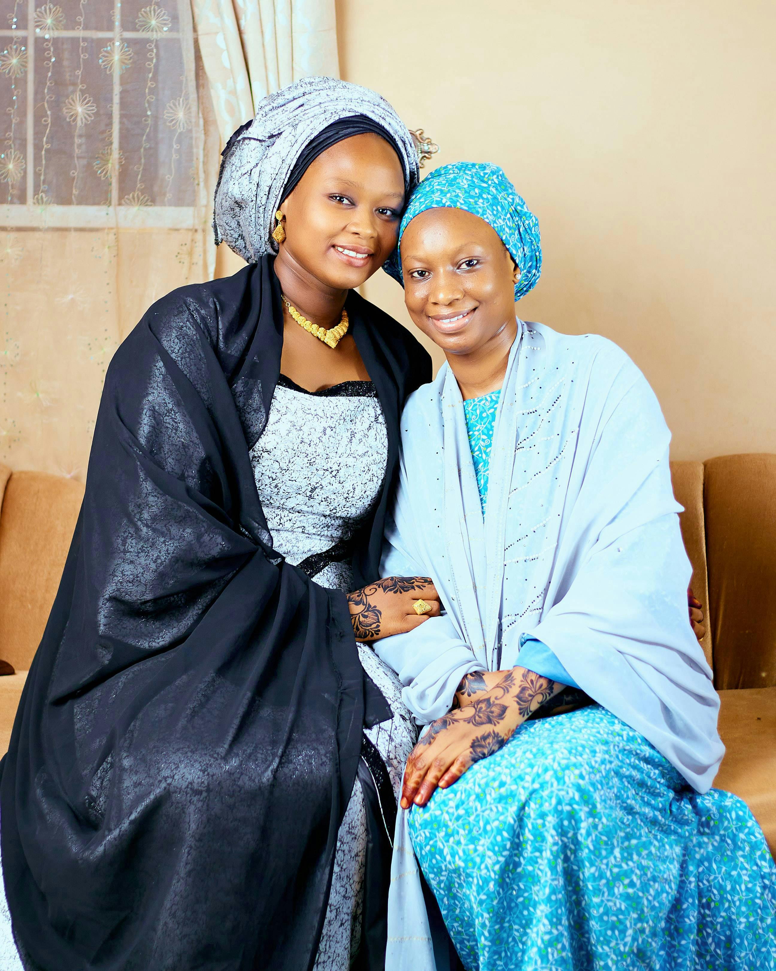 Two women in beautiful traditional African attire posing indoors with elegant smiles.