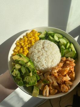 A vibrant poke bowl featuring rice, avocado, corn, cucumber, and chicken in natural light.