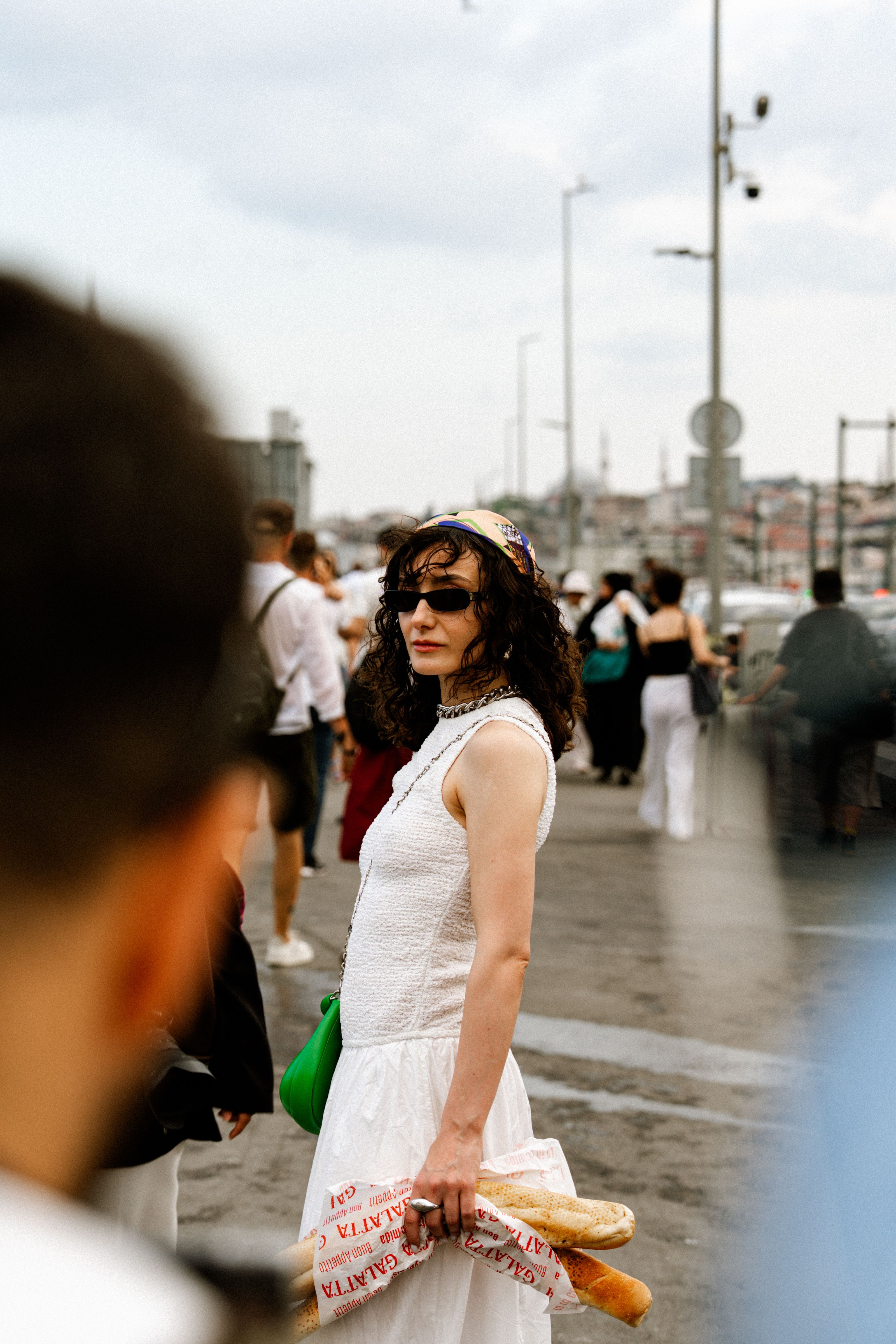 Mulher de vestido branco carregando baguetes em uma movimentada cena de rua da cidade.