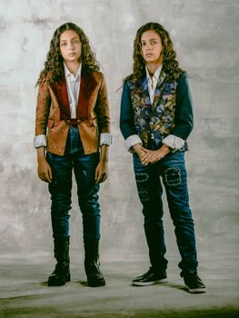 Two teenage twins pose in trendy jackets against a textured backdrop, exuding youthful style and confidence.