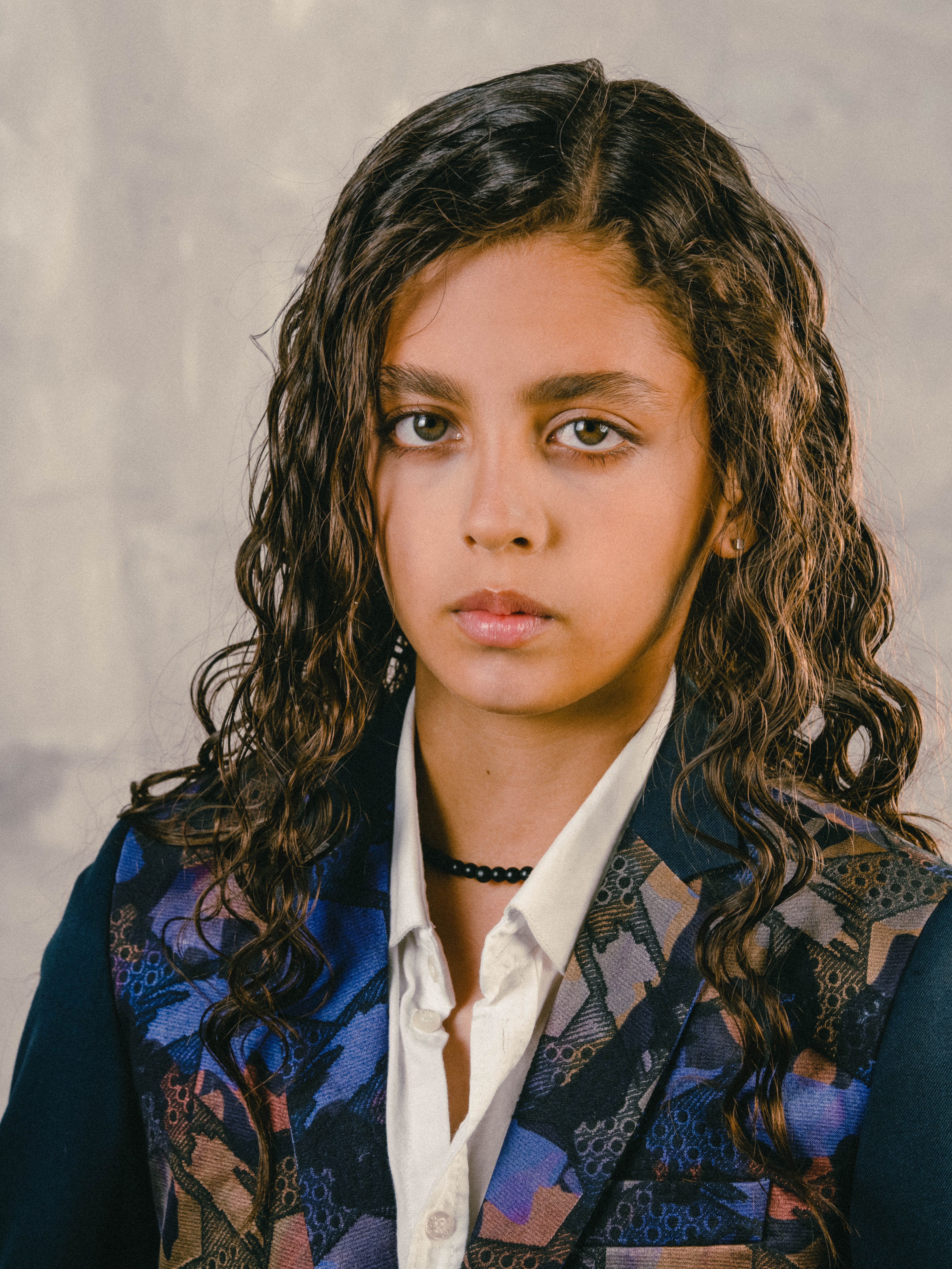 A thoughtful young girl with curly hair wearing a blazer and shirt, posing indoors.