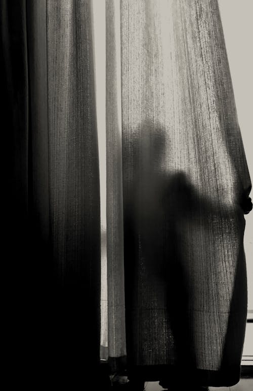 Grayscale Photography of Person Standing While Hiding on Curtain