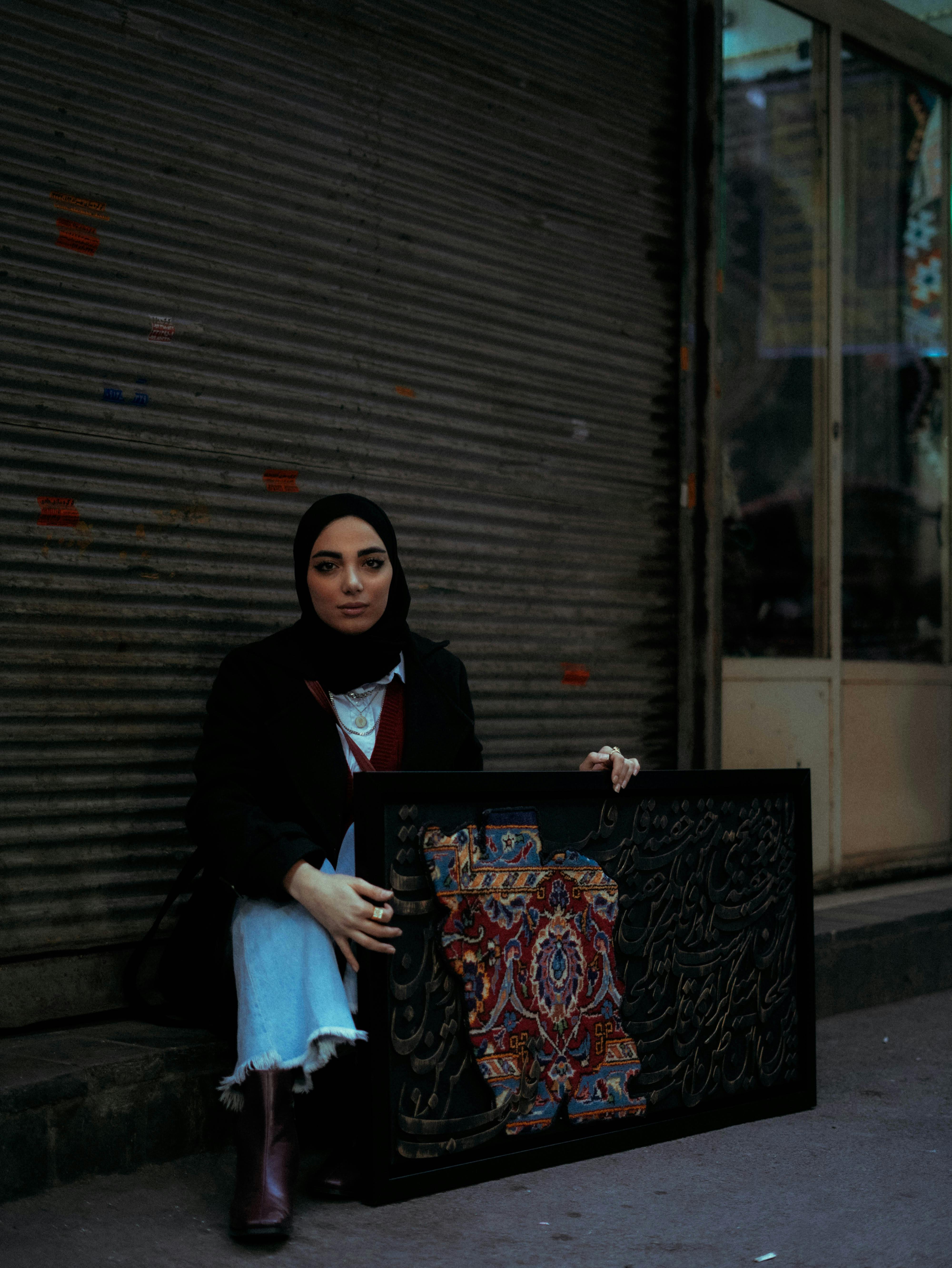 Young woman in hijab sitting outdoors holding an intricately designed textile art piece framed in black.