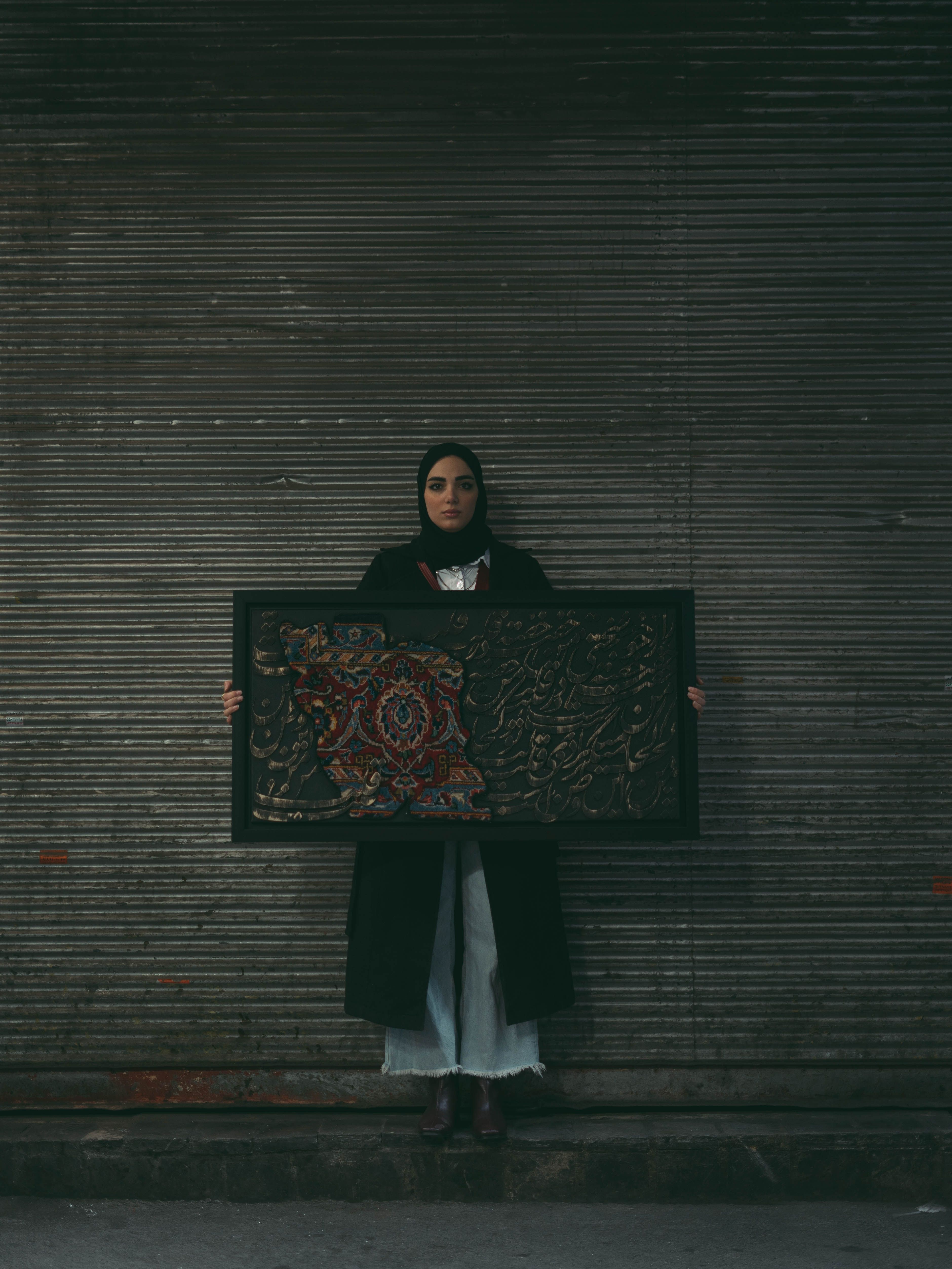 Middle Eastern woman holding an ornate artwork, standing against a corrugated metal backdrop.