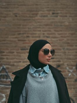 Fashionable woman wearing hijab and sunglasses posing in an urban setting with a brick wall background.