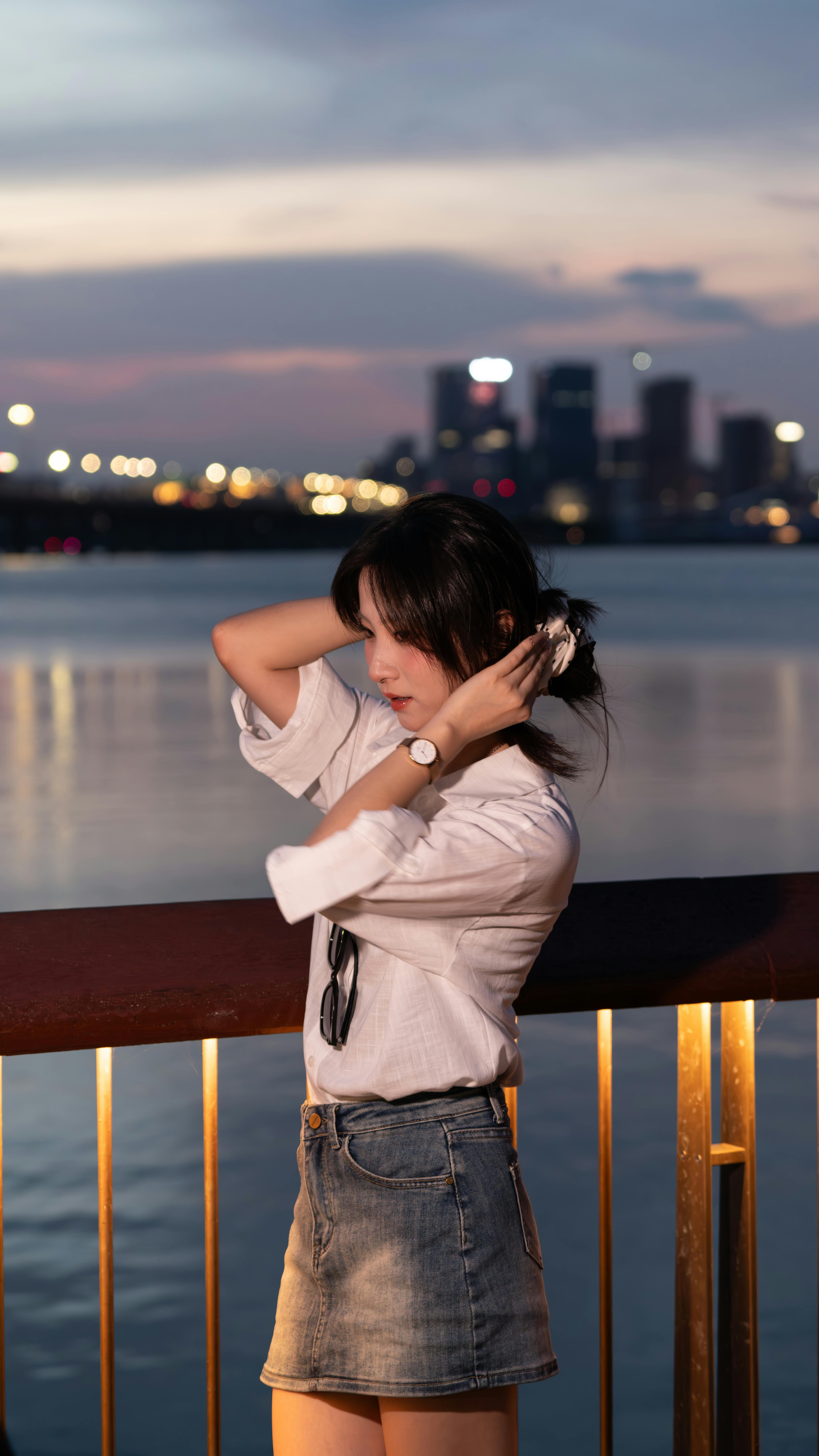A young woman in casual attire enjoys a peaceful sunset by the riverside with a cityscape backdrop.
