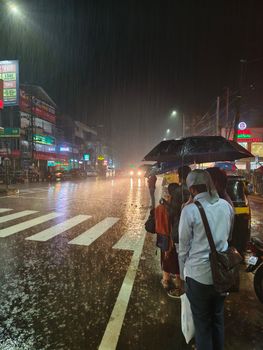People with umbrellas wait at a crosswalk during heavy rain on a busy street at night.