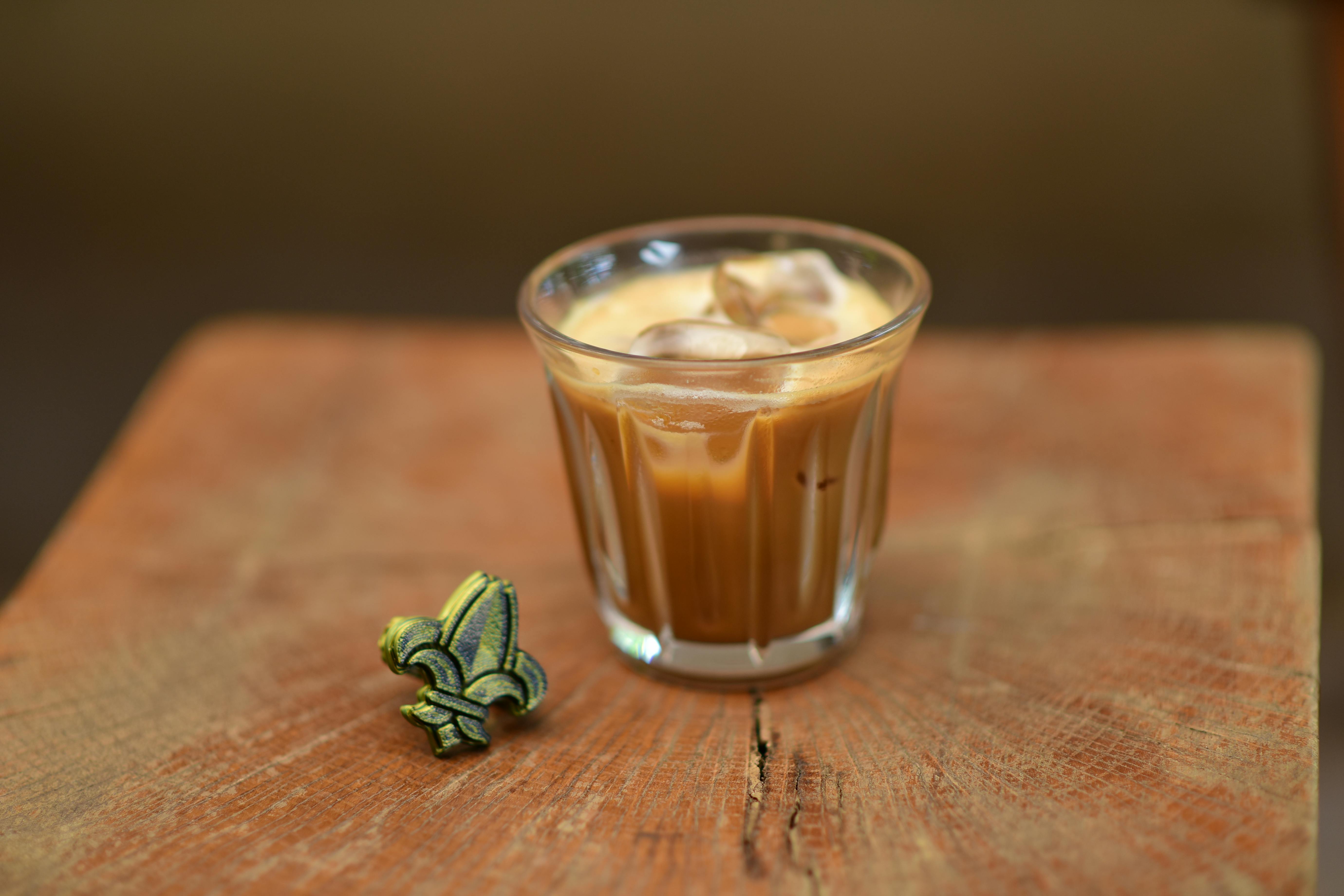 Glass of iced coffee with ice cubes beside a decorative fleur-de-lis on a rustic wooden table. Does Drinking Coffee Cause Acid Reflux?