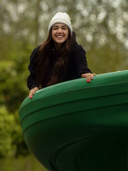 Happy young woman with long hair and beanie plays on a large green structure in a park.
