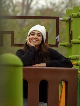 A young woman wearing a white beanie smiles warmly outdoors, leaning on a colorful structure.