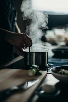 A person stirring a steaming pot on an electric stove with kitchenware spread out.