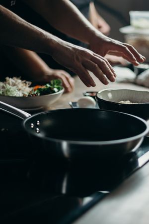 Hands seasoning food beside a pan on a dark stove. Photo by cottonbro studio on Pexels.