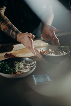 Tattooed chef presenting Asian cuisine with fresh ingredients in a modern kitchen setting.