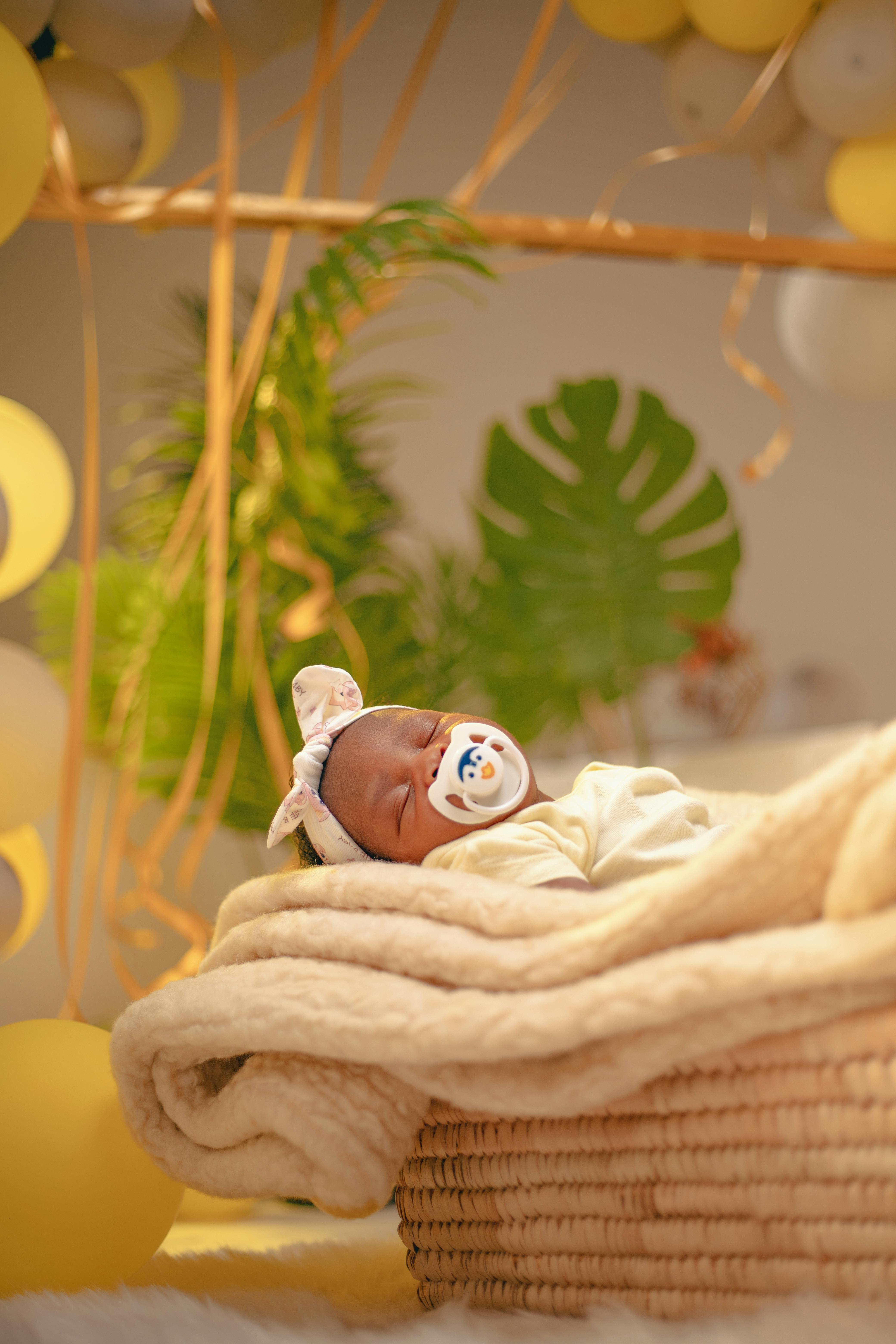 бесплатная Новорожденный мирно покоится в плетеной корзине, окруженной желтыми воздушными шарами и зеленью. Стоковое фото