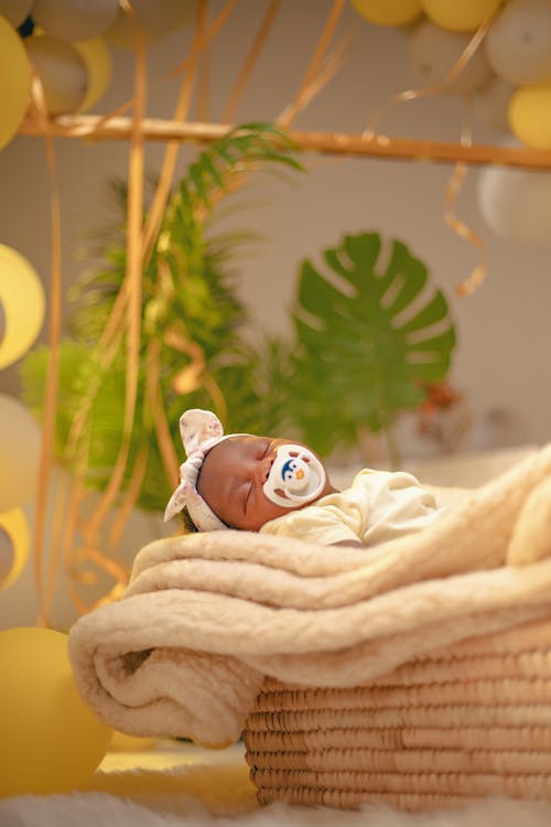 бесплатная Новорожденный мирно покоится в плетеной корзине, окруженной желтыми воздушными шарами и зеленью. Стоковое фото