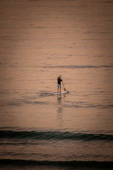 A lone paddleboarder enjoys a peaceful ride on the ocean during sunset, surrounded by warm orange hues.