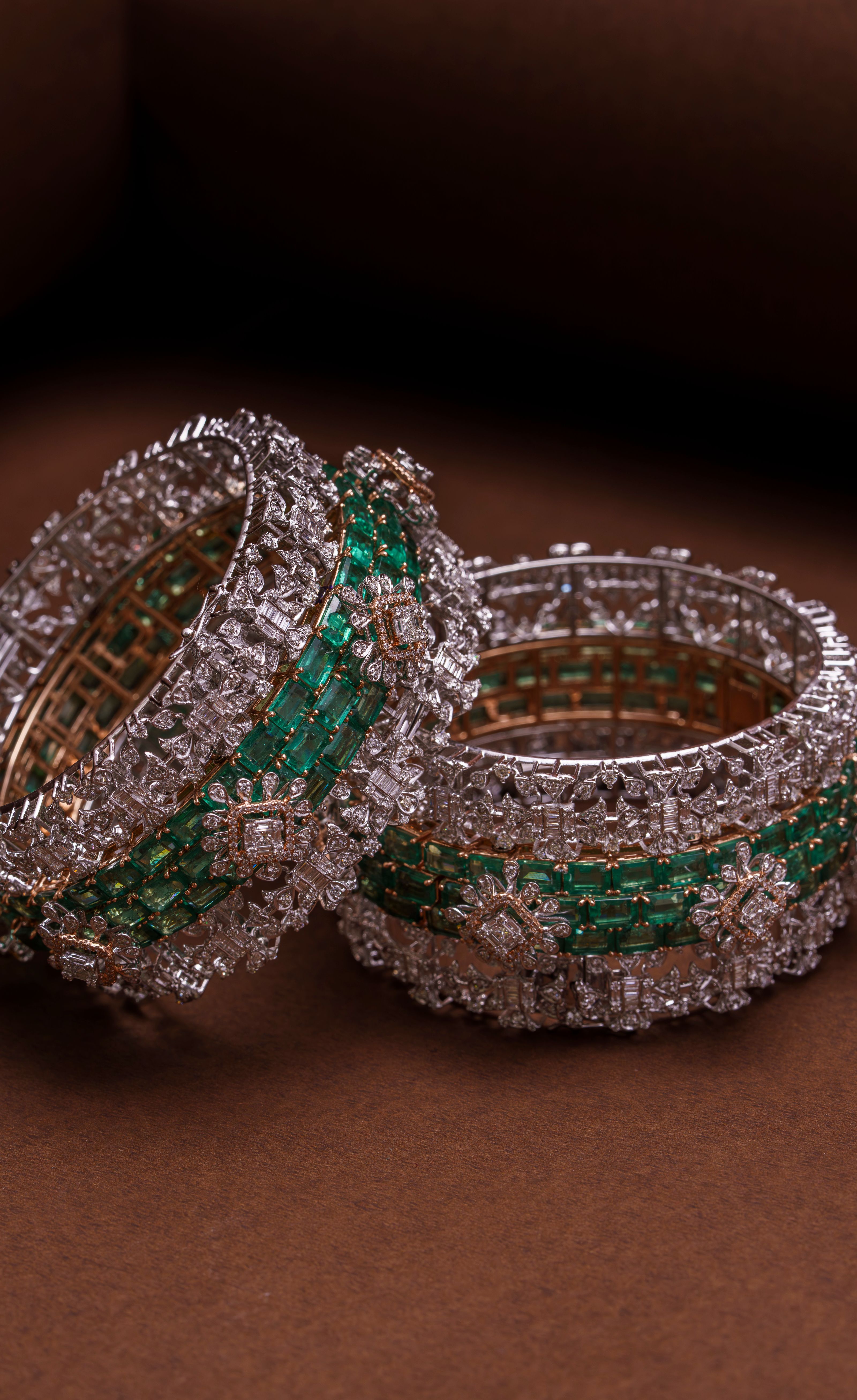 Pulseiras luxuosas incrustadas de esmeraldas e diamantes em um fundo marrom chocolate, exibindo um artesanato complexo.