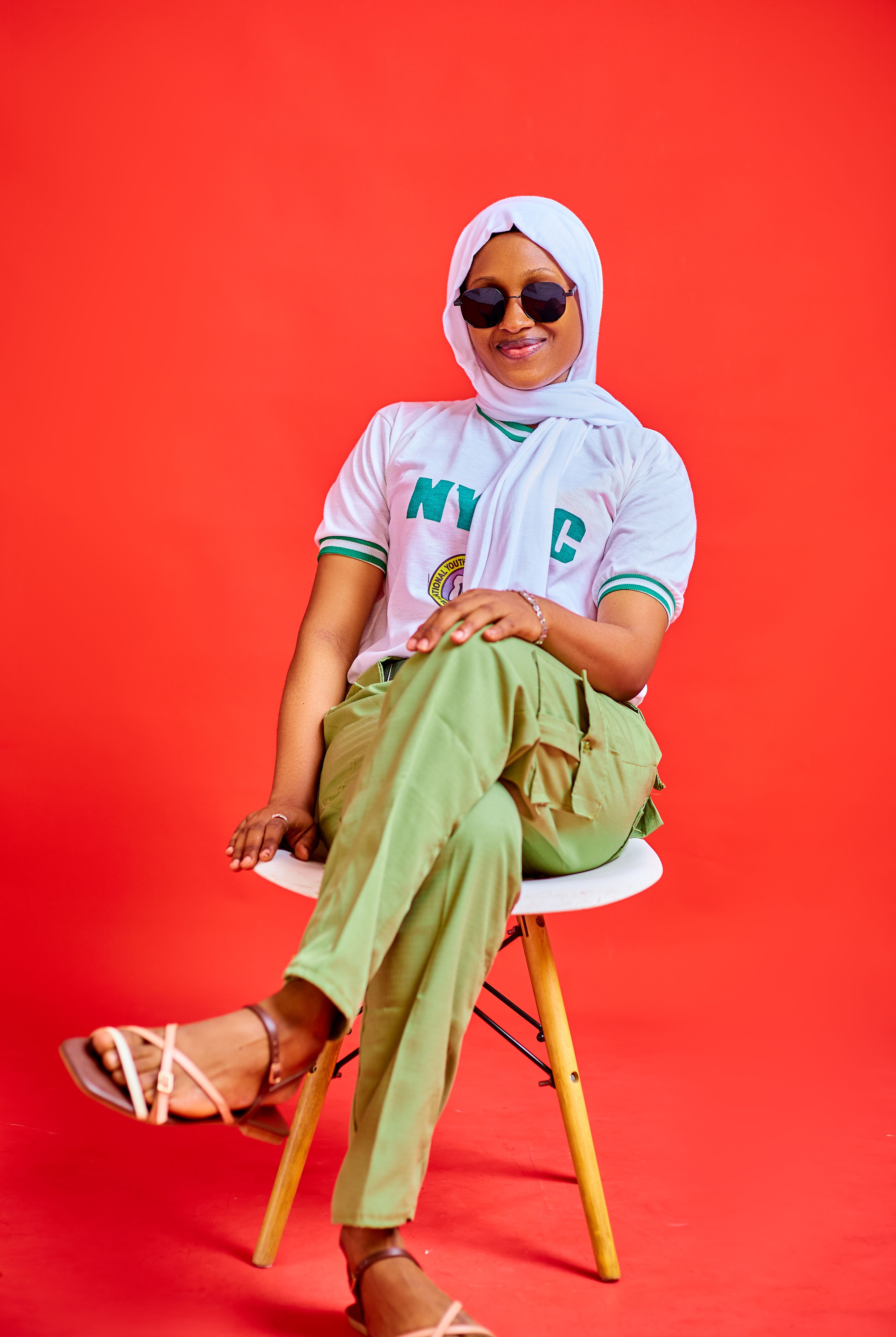 Portrait of a Nigerian woman in traditional NYSC attire sitting against a red background.