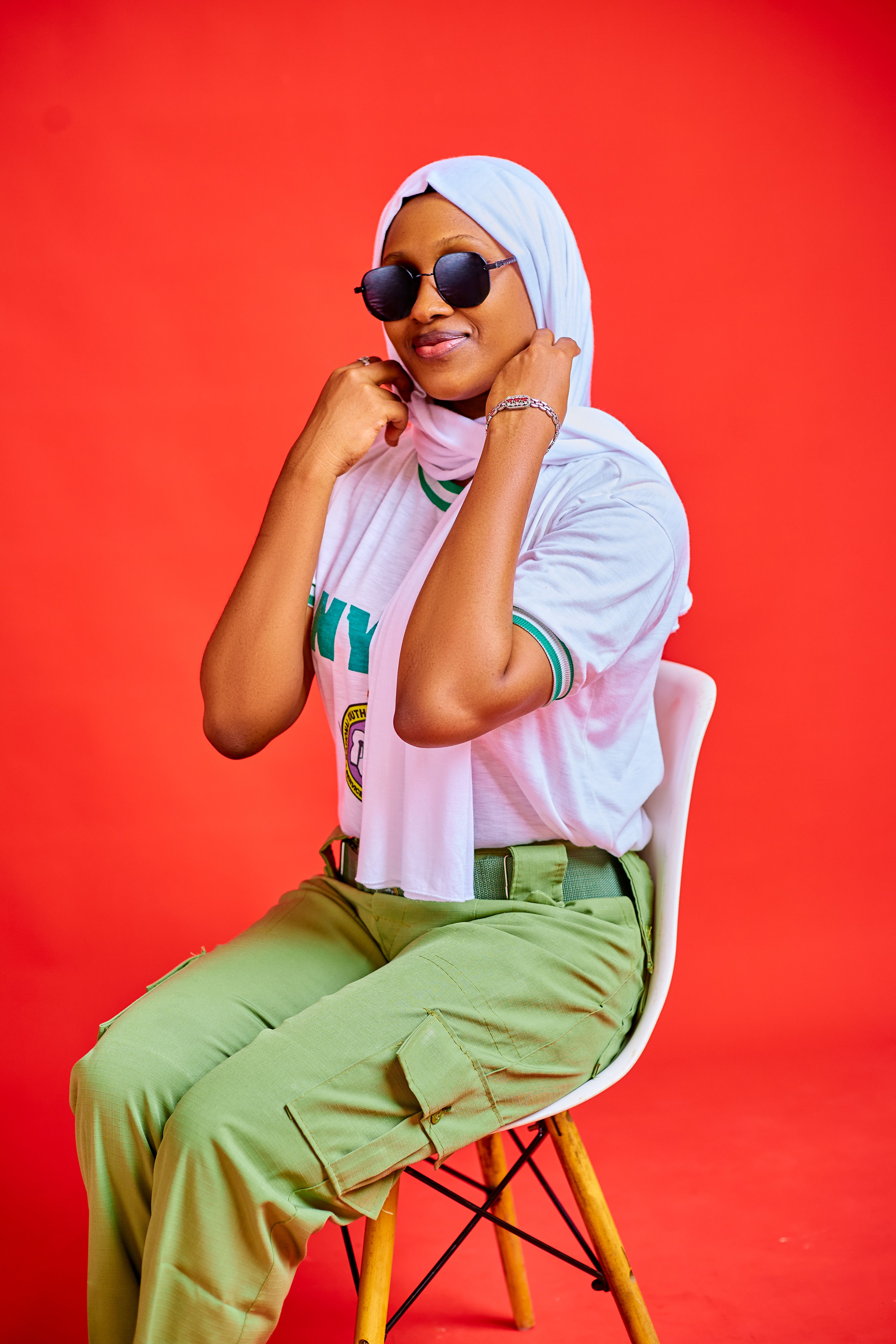 Fashionable woman in sunglasses and hijab, sitting against a bold red backdrop.