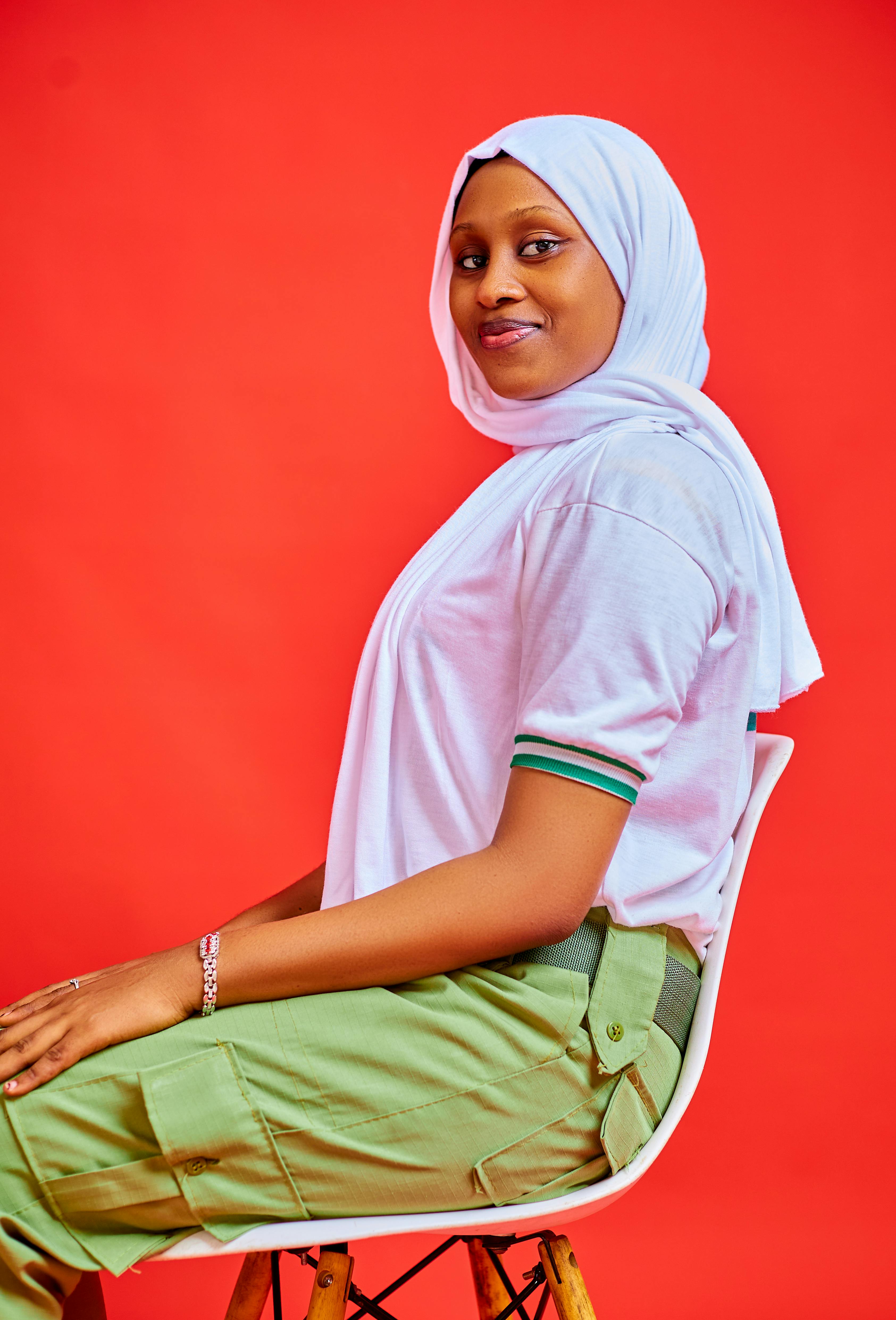 Confident woman in white hijab sitting against vibrant red background, smiling warmly.
