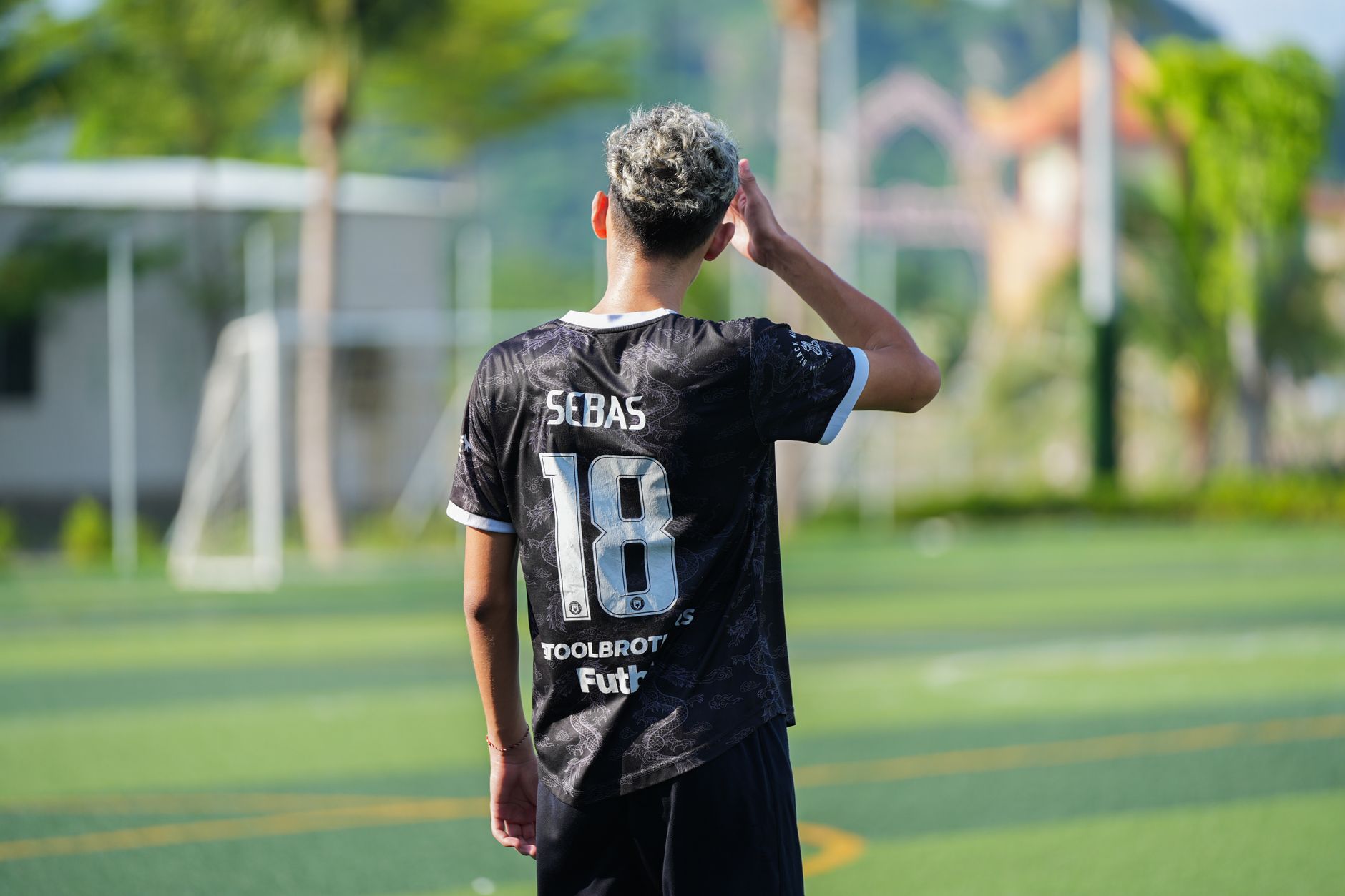 Young player in black jersey on an outdoor soccer field during the day, facing away from the camera.