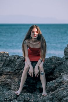 Confident woman in alternative fashion posing on rocky shore with ocean backdrop.