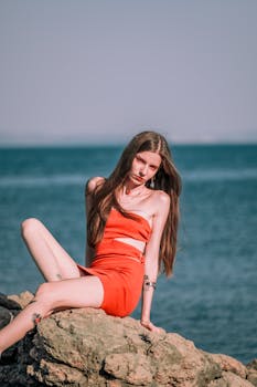 Model in orange dress on rocky shore with ocean backdrop, exuding confidence.