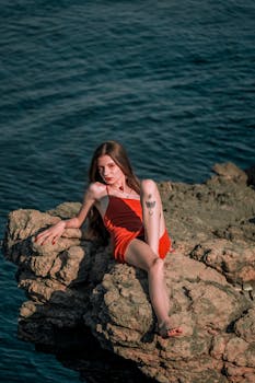 A fierce young woman in a red summer outfit poses dramatically on sea rocks.