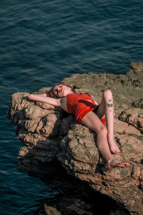 Free Confident model in red dress posing dramatically on rocky seaside cliff under bright sunlight. Stock Photo