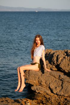 A striking seaside portrait of a woman sitting on rocky beach enjoying the sunset.