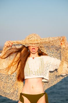 A playful young woman in crochet fashion poses humorously by the seaside, embodying summer vibes.