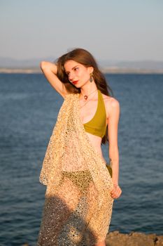 Model in green bikini and sheer shawl on a beach during sunset. Fashion editorial photo.