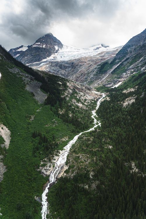 Free Majestic aerial scenery capturing a mountain valley with a winding stream and glacier under dramatic skies. Stock Photo