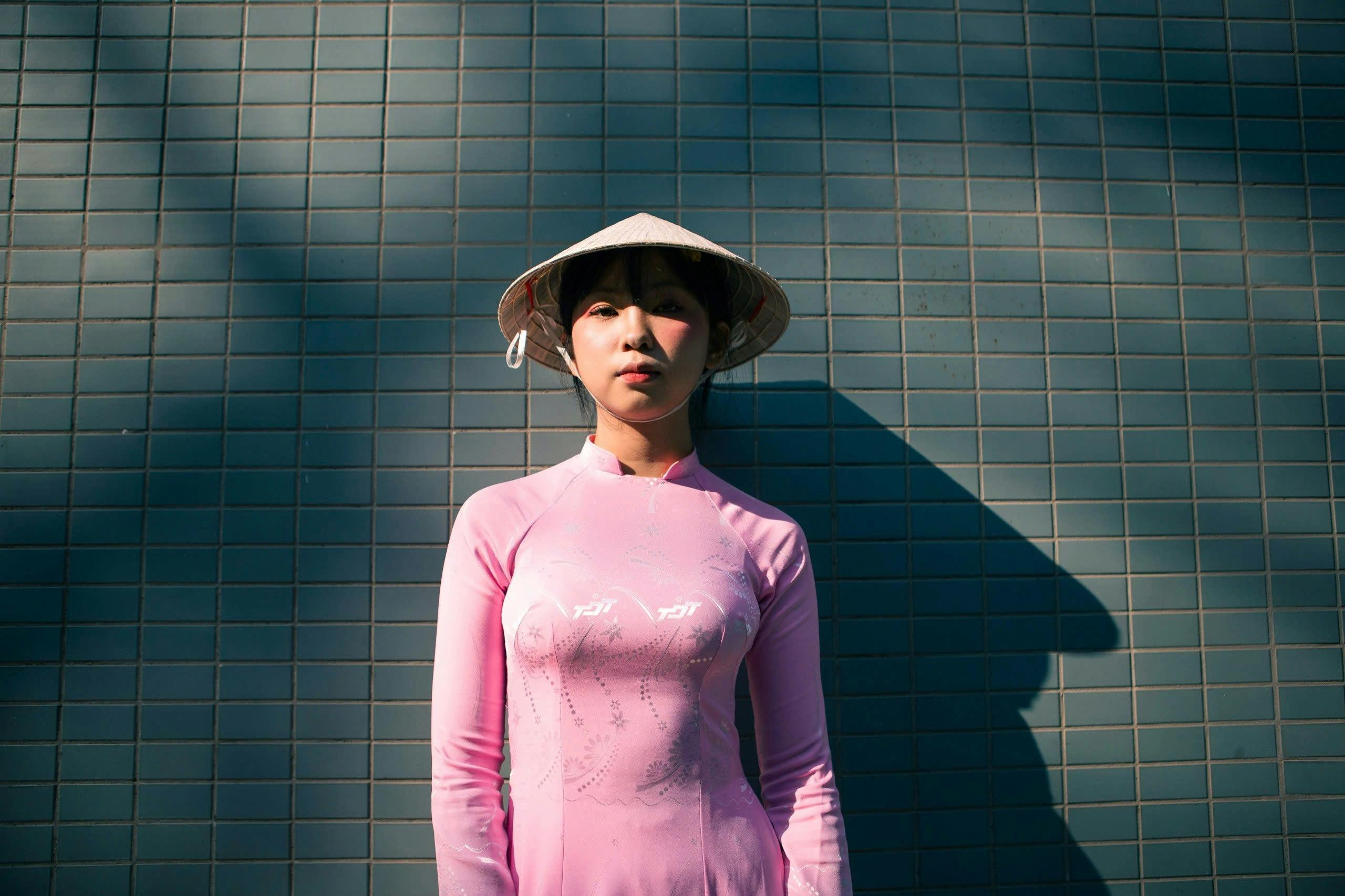 Woman wearing pink ao dai and conical hat stands against a tiled wall.