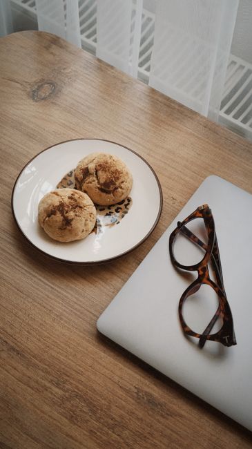 A cozy workspace setup with cookies, a laptop, and reading glasses on a wooden desk.