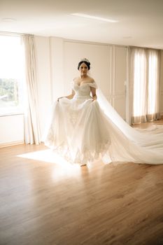 A bride in a flowing gown stands elegantly in a sunlit room, exuding grace and beauty.