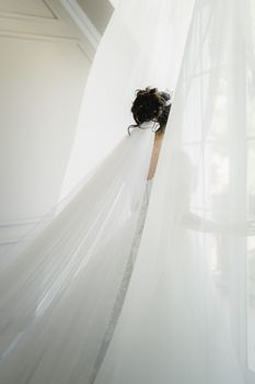 A bride in an elegant white gown stands by a window, captured in a soft and bright ambiance.