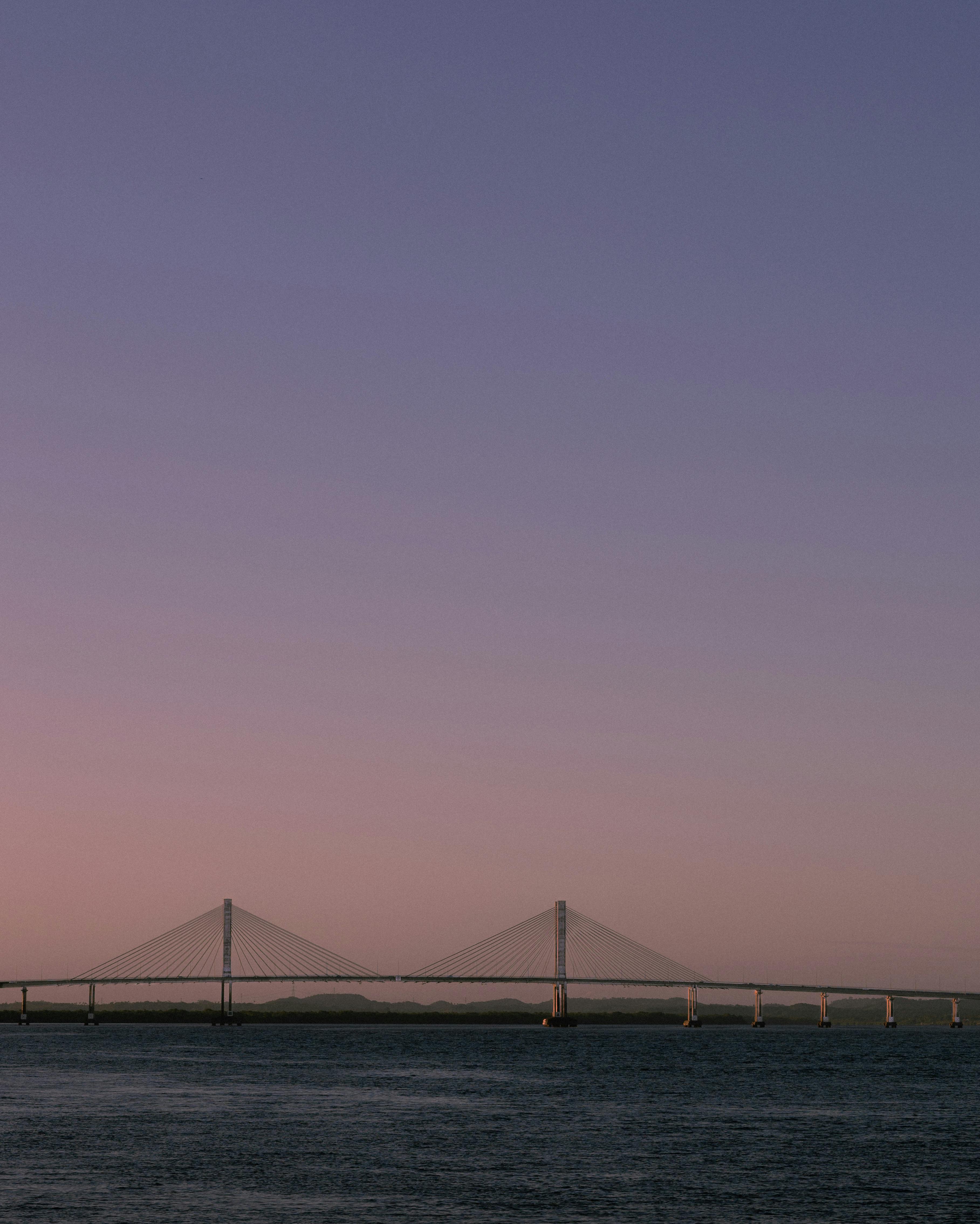 Serene sunset view of a bridge silhouette stretching over calm water reflecting purple hues.