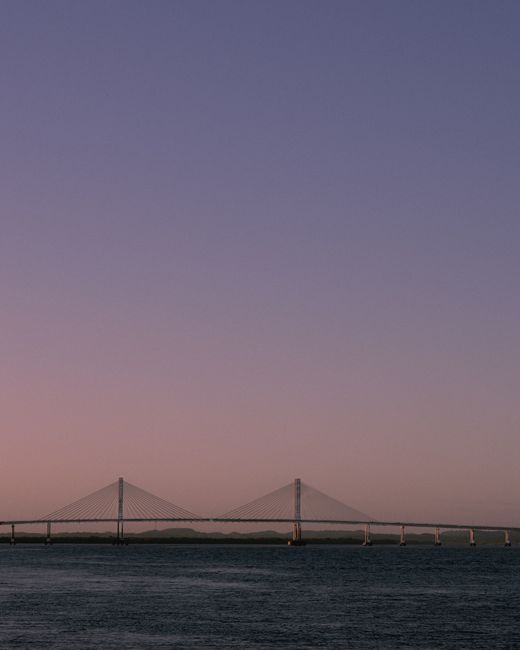 Serene sunset view of a bridge silhouette stretching over calm water reflecting purple hues.
