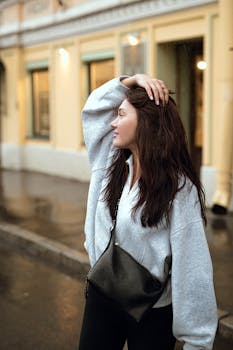 A thoughtful young woman stands outdoors on a rainy city street, enjoying the moment.