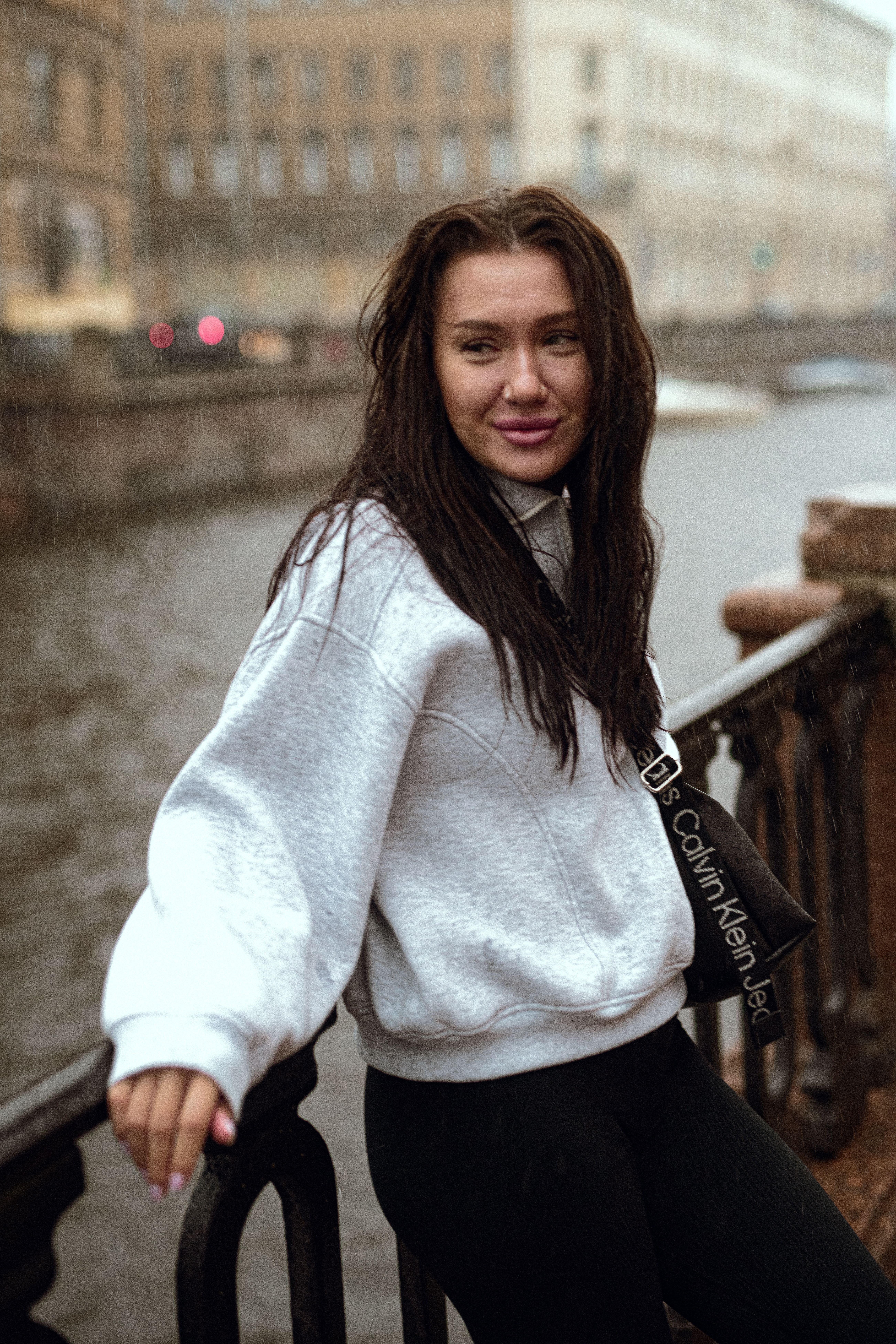 A woman in a casual outfit poses by a riverside railing on a cloudy autumn day.